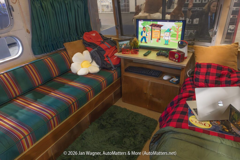 Interior of a cozy RV with a striped couch, flower pillow, backpack, TV playing a cartoon, small desk, red plaid bedding, laptop, and books.