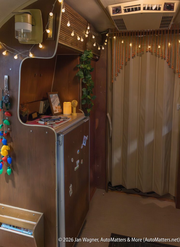 Interior of a retro camper van featuring wood paneling, a small fridge, string lights, decorative beads, and various personal items.