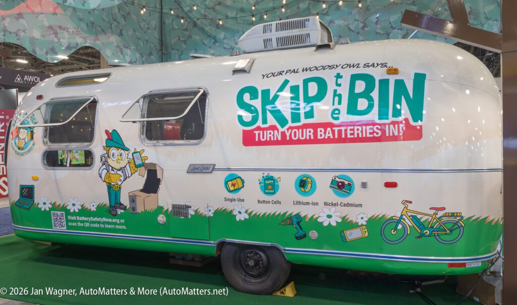 A silver trailer displays a cartoon owl with a battery recycling message: "Skip the Bin, Turn Your Batteries In!" and lists battery types, beside a green bike illustration.
