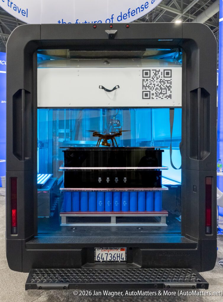 Open rear view of a high-tech vehicle displaying a shelving unit with blue cylindrical containers, a small drone, and a visible QR code.