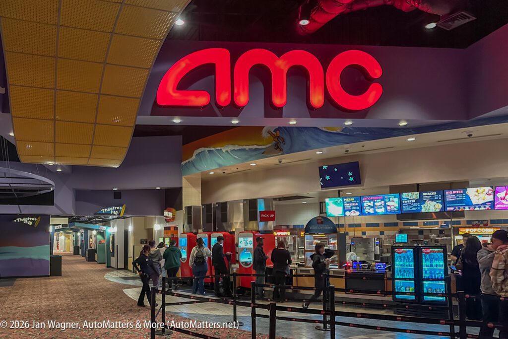 AMC movie theater lobby with people standing at the concession stand, offering snacks and drinks under bright lighting.