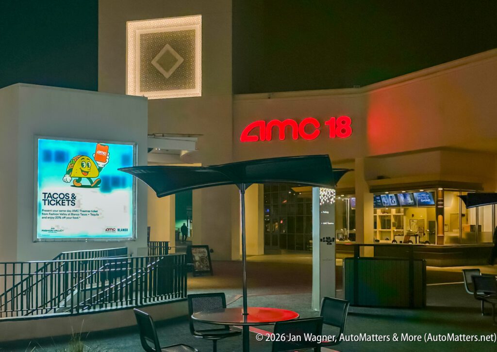 Outdoor view of an AMC 18 movie theater entrance at night, featuring a large illuminated billboard and empty café tables in the foreground.