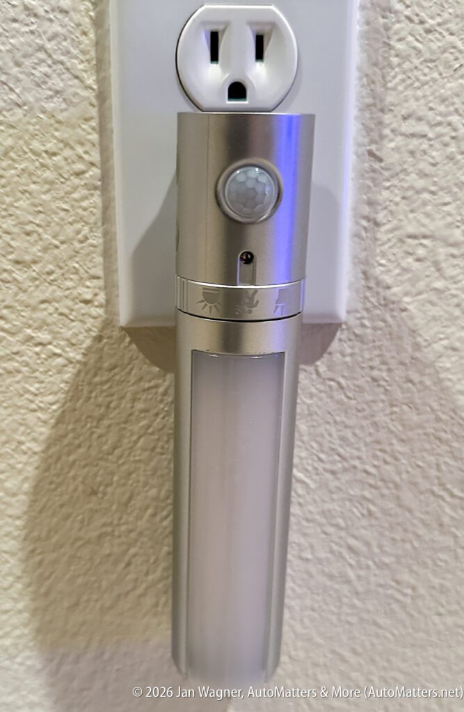 A silver motion-sensor night light is plugged into a wall outlet against a textured beige wall.