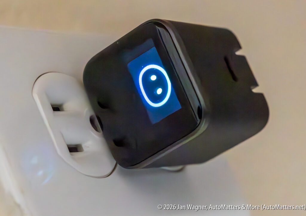 A black smart plug with a digital display showing a lit blue smiley face icon is plugged into a white electrical outlet.