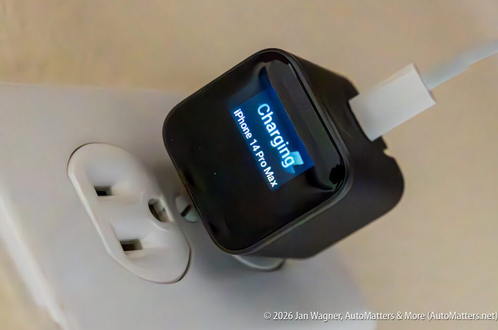 A black charger plugged into a wall outlet displays "Charging iPhone 14 Pro Max" on its screen, with a white charging cable connected.