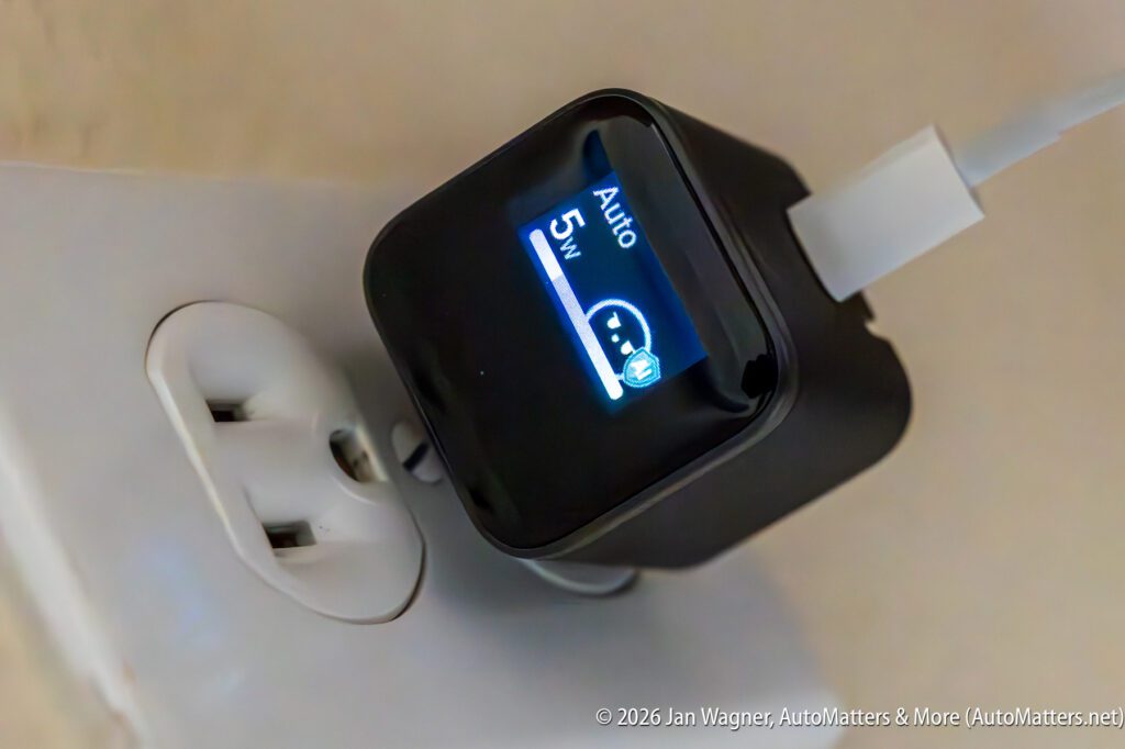 A black USB charger with a digital display shows "5W" while plugged into a wall outlet, charging a device via a white cable.