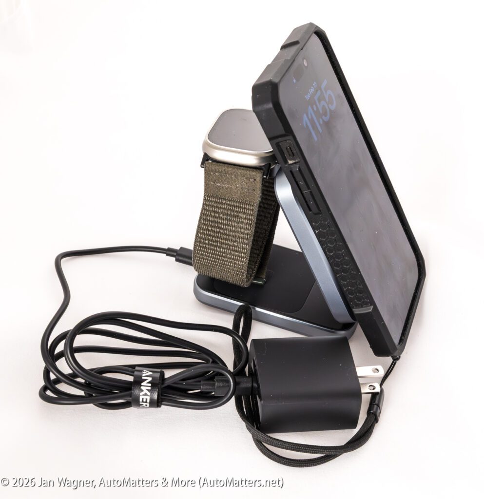 A smartphone and smartwatch rest on a charging stand, with a black USB wall adapter and cable attached to the stand.