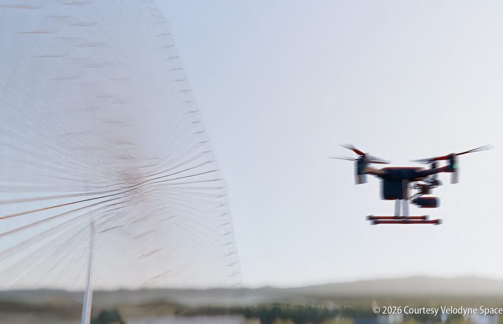 A drone flies outdoors near a large parabolic antenna, with a blurry landscape and sky in the background.
