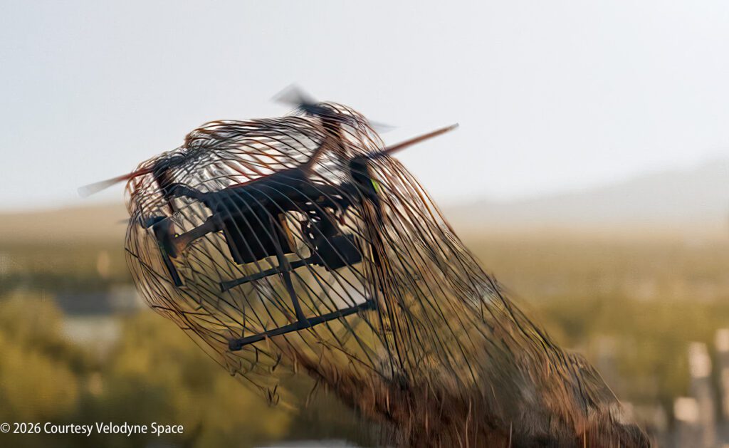 A drone is ensnared in a net outdoors, with blurred landscape in the background. ©2026 Courtesy Velodyne Space.