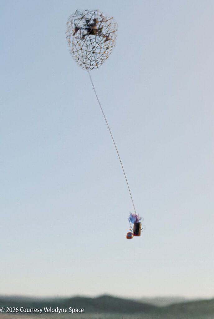 A small drone is captured mid-air by a net attached to a long tether, with hills and a clear sky in the background.