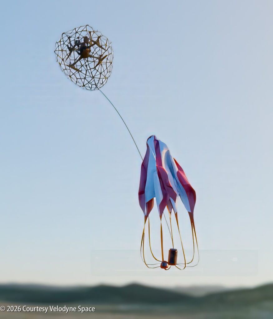 A small aerial device with a netted sphere, trailing a white and red ribbon tail, is suspended in midair against a clear sky.