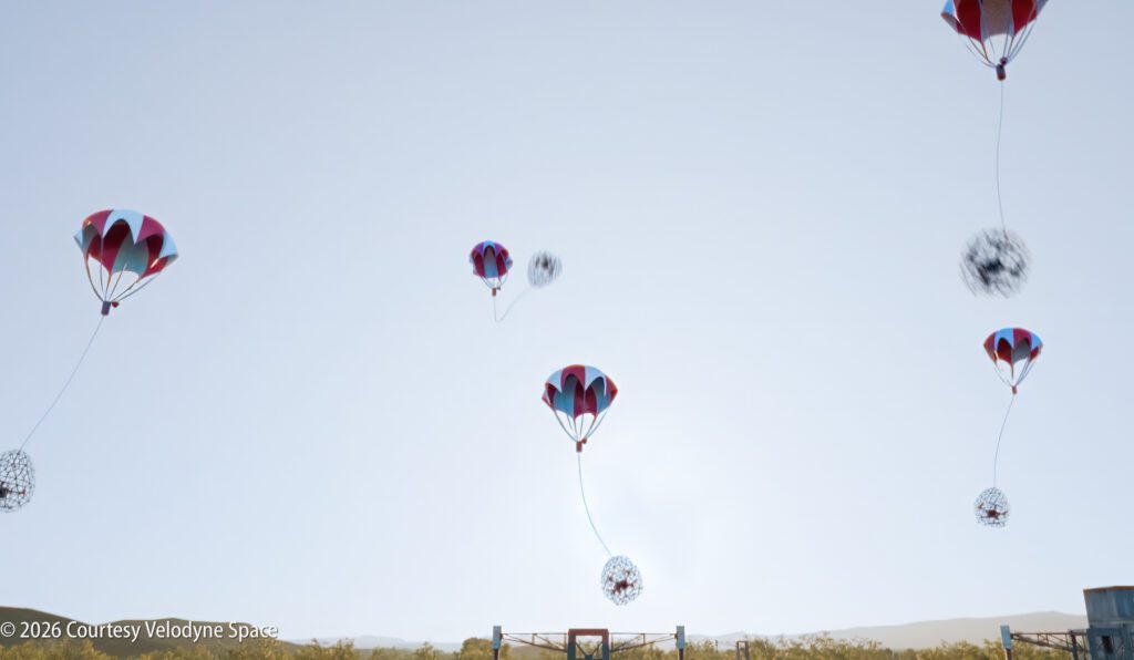 Six spherical objects attached to red and blue parachutes descend through the sky over a grassy landscape, with a metal structure visible below.
