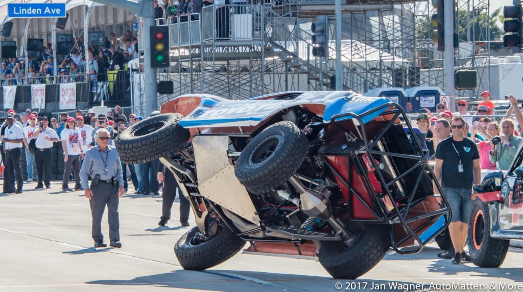 A race truck driving on two side wheels during a motorsport event, surrounded by officials and spectators.