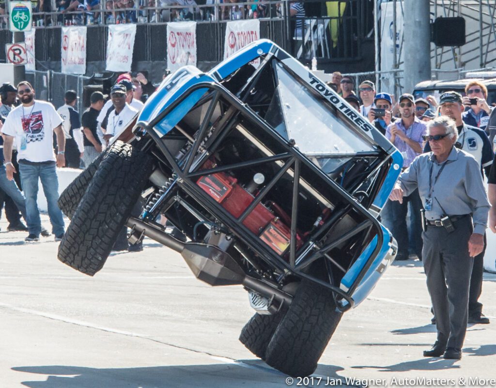 A blue off-road truck drives on two wheels at an event, with a crowd watching and a man standing nearby.