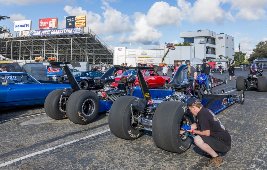 A person works on a dragster in the pit area of a racetrack, surrounded by other race cars and people, with grandstands and a building in the background.