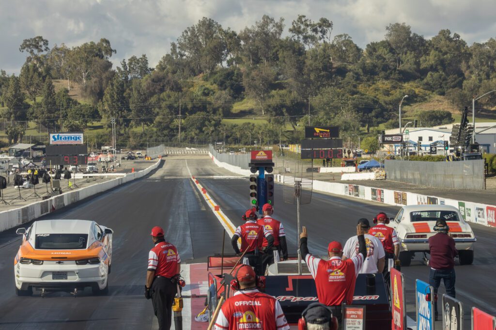 Two cars line up at the start of a drag race as officials in red uniforms oversee the event. The track is surrounded by trees and various buildings.
