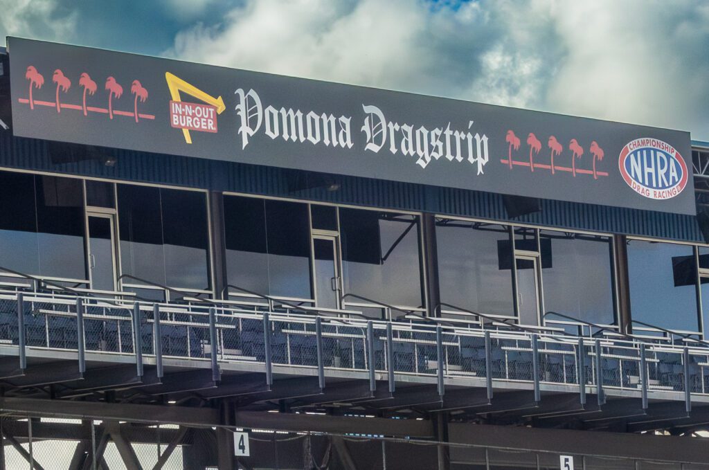 A sign for Pomona Dragstrip with logos for In-N-Out Burger and NHRA is displayed above seating and glass windows under a cloudy sky.