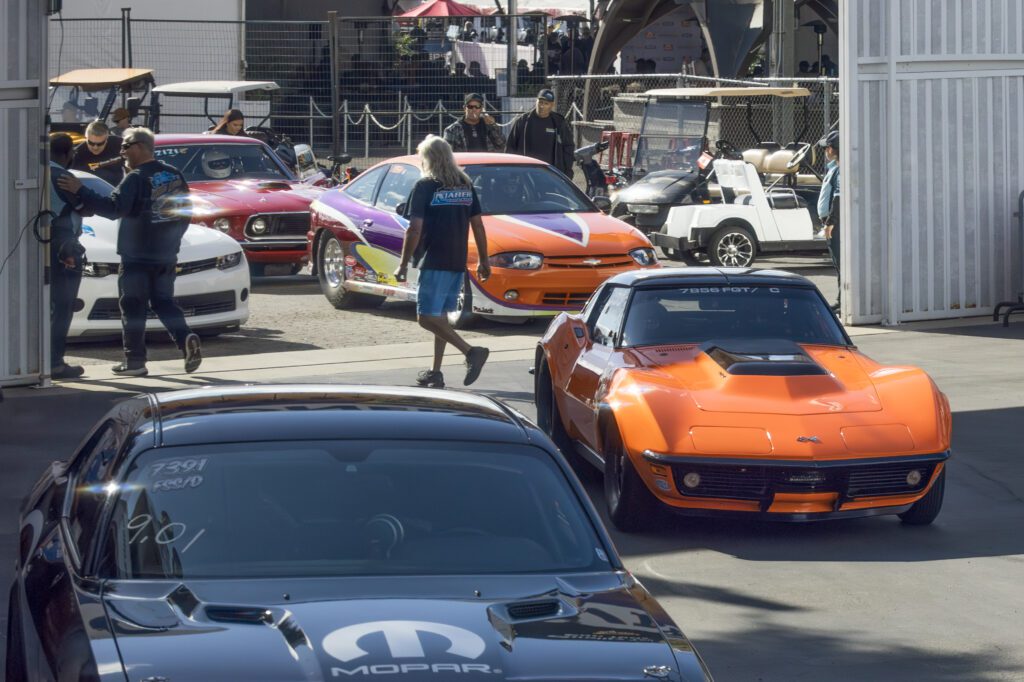 Several classic and modern cars, including an orange sports car in the foreground, are parked at an outdoor car event with people walking around.