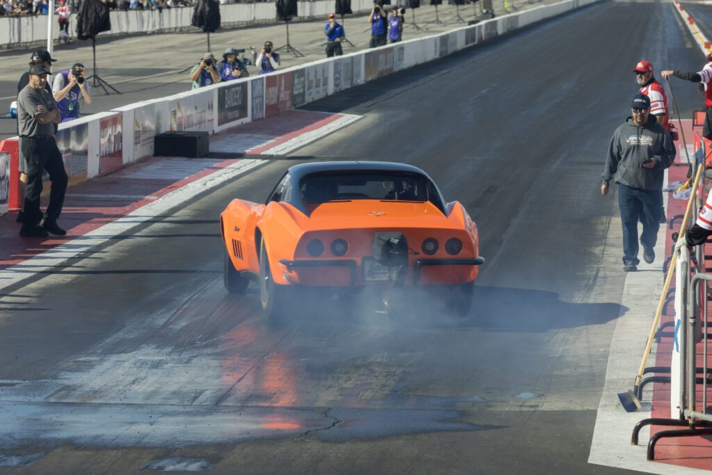 An orange race car accelerates on a drag strip, producing smoke from its tires as officials and spectators observe from the sidelines.