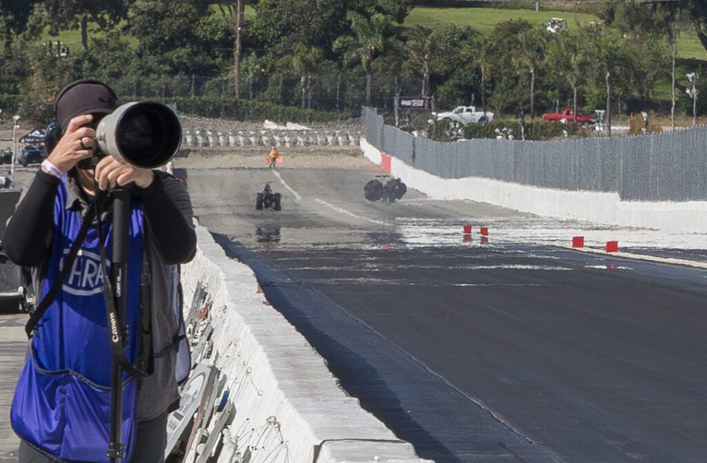 A photographer with a large camera stands at the edge of a race track, capturing images; two vehicles and barriers are visible in the distance.