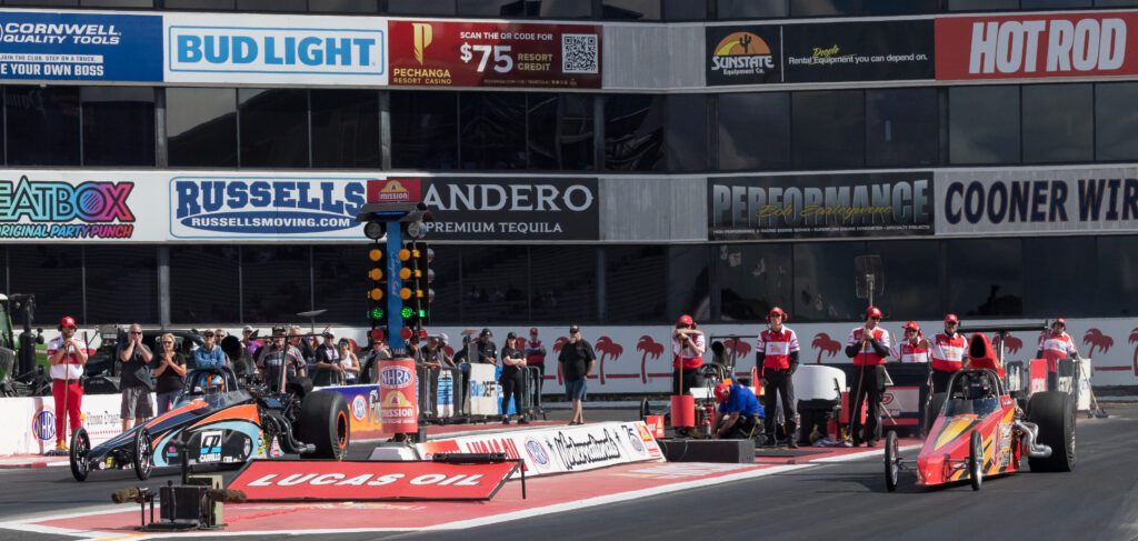 Two drag racing cars at the starting line of a racetrack, with crew members and officials standing nearby and sponsor banners in the background.