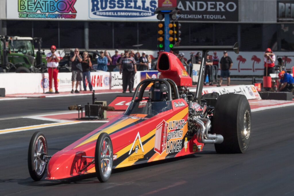 A dragster car with sponsor logos is on a drag racing track, preparing to start the race as spectators watch and lights signal at the starting line.