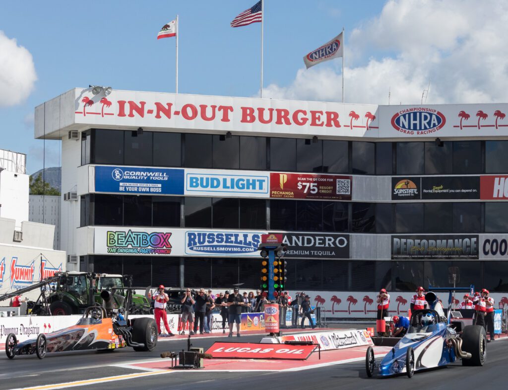 Two drag racing cars at the starting line of an NHRA event, with the In-N-Out Burger building and flags in the background.