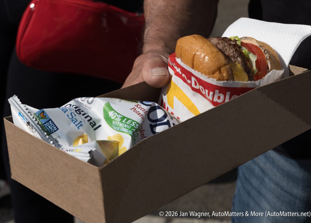 A person holds a cardboard tray containing a wrapped In-N-Out burger and a bag of Kettle Brand potato chips.
