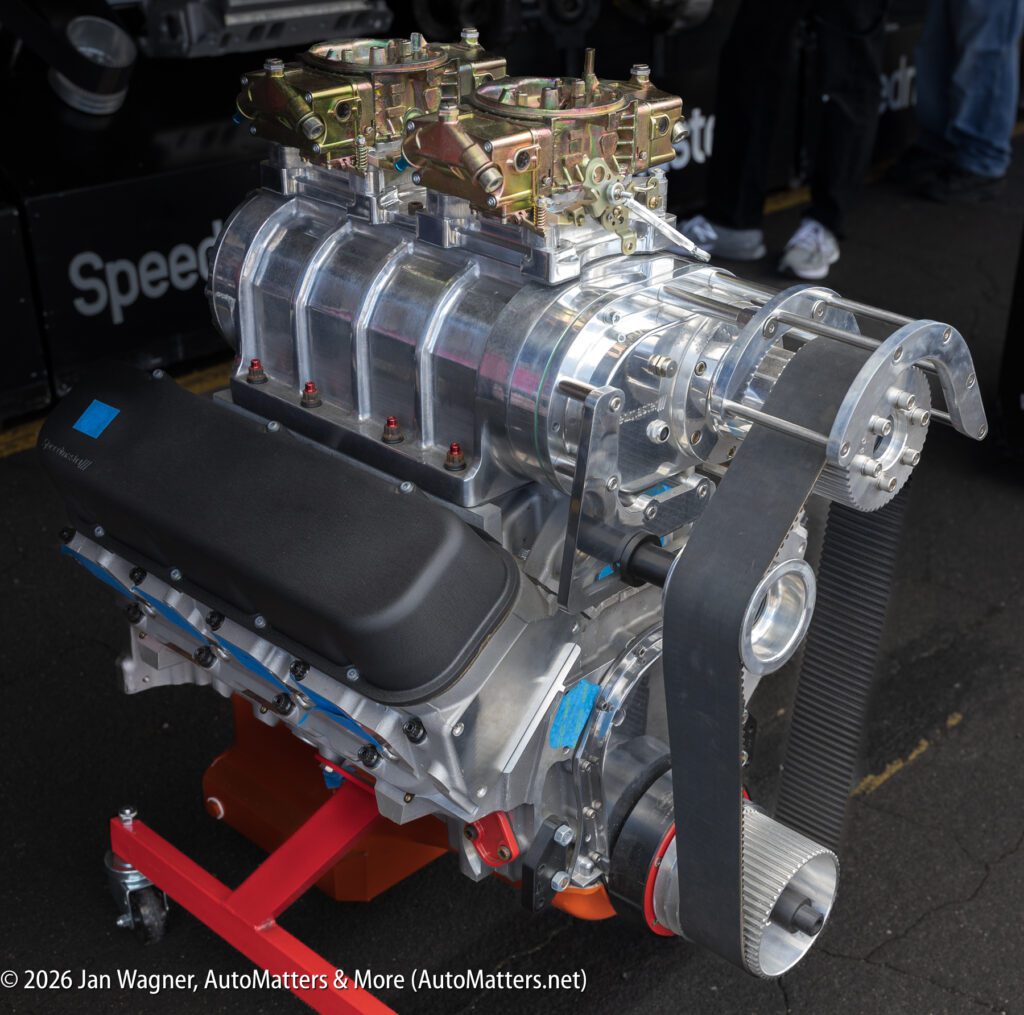 A high-performance supercharged V8 engine with dual carburetors on display, featuring exposed pulleys and belt drive components.