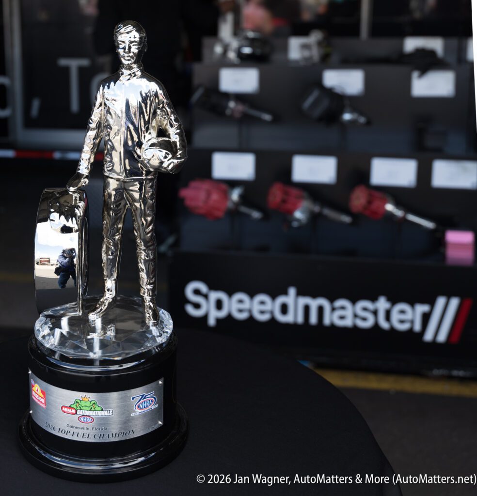 A silver racing trophy with a driver figure stands on a table; Speedmaster equipment is displayed in the background.