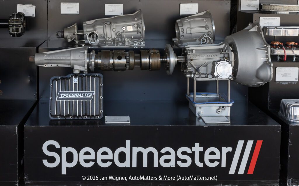 Display of automotive transmission components and a Speedmaster-branded electronic control unit, arranged on a black stand labeled "Speedmaster" at an exhibition.