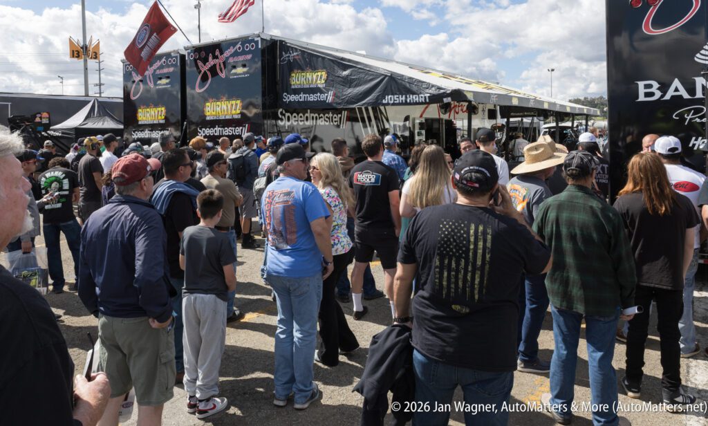 Crowd of people standing in line outdoors at an event near racing trailers and sponsor banners under a partly cloudy sky.