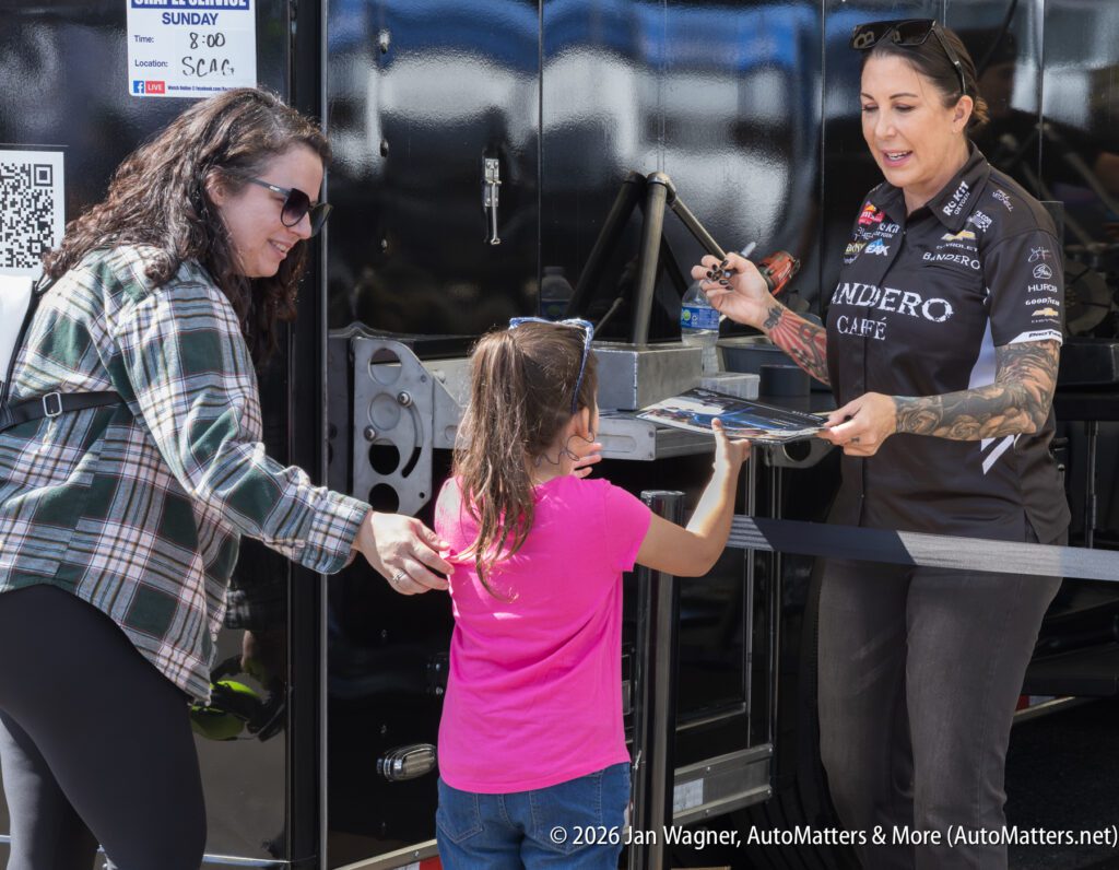 A woman hands a poster to a young girl while another woman stands beside her, outdoors near a racing team trailer.