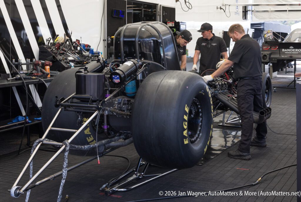 Three mechanics work on a drag racing car in a garage, with the rear wheels exposed and various tools and equipment visible around them.