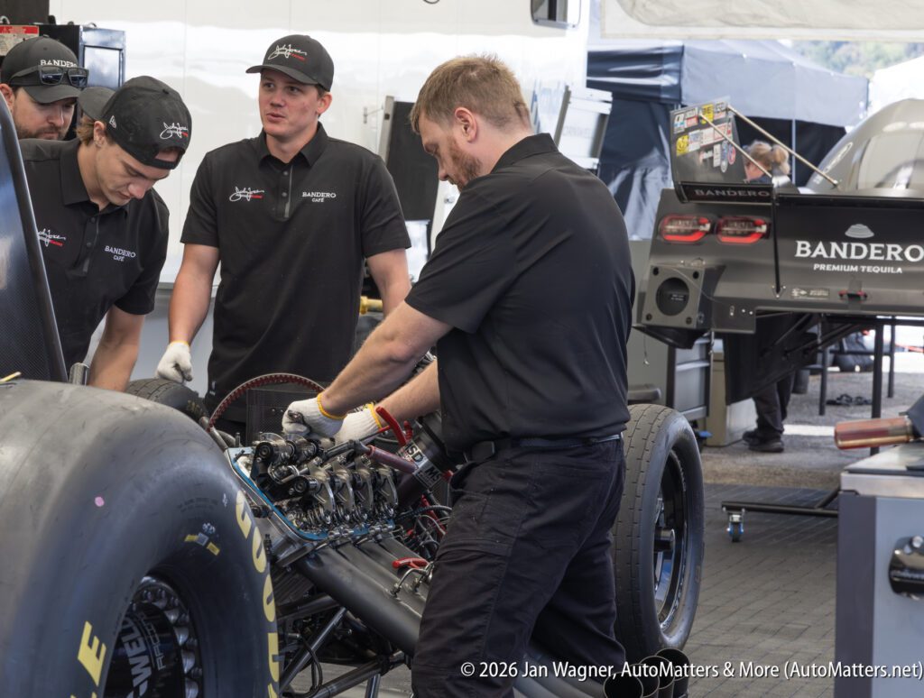Four team members in black shirts work on the engine of a race car in a garage area, with tools and car parts visible around them.