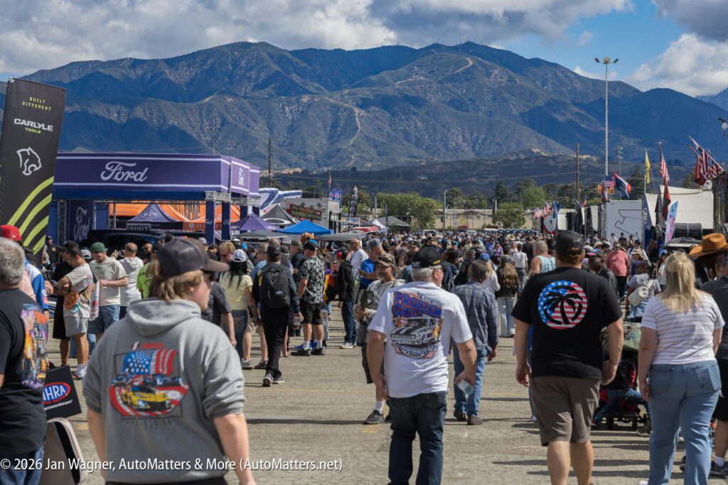A large crowd walks through an outdoor event with vendor booths and mountains in the background under a partly cloudy sky.