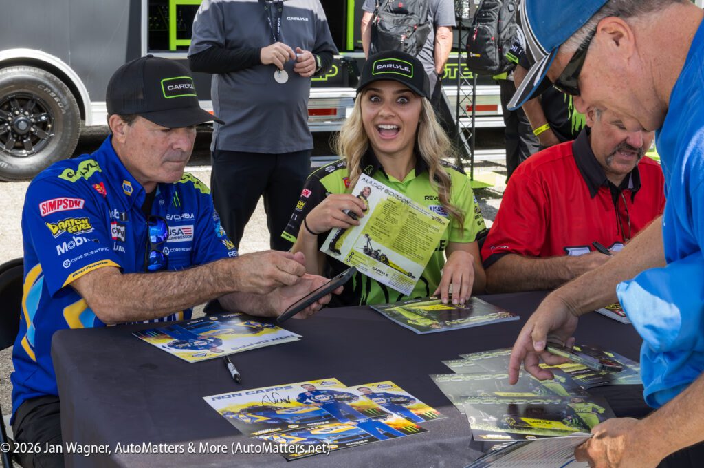 Two motorsports team members sign autographs at an outdoor event. The woman in the center smiles while holding a signed photo. Others wait and watch at the table.