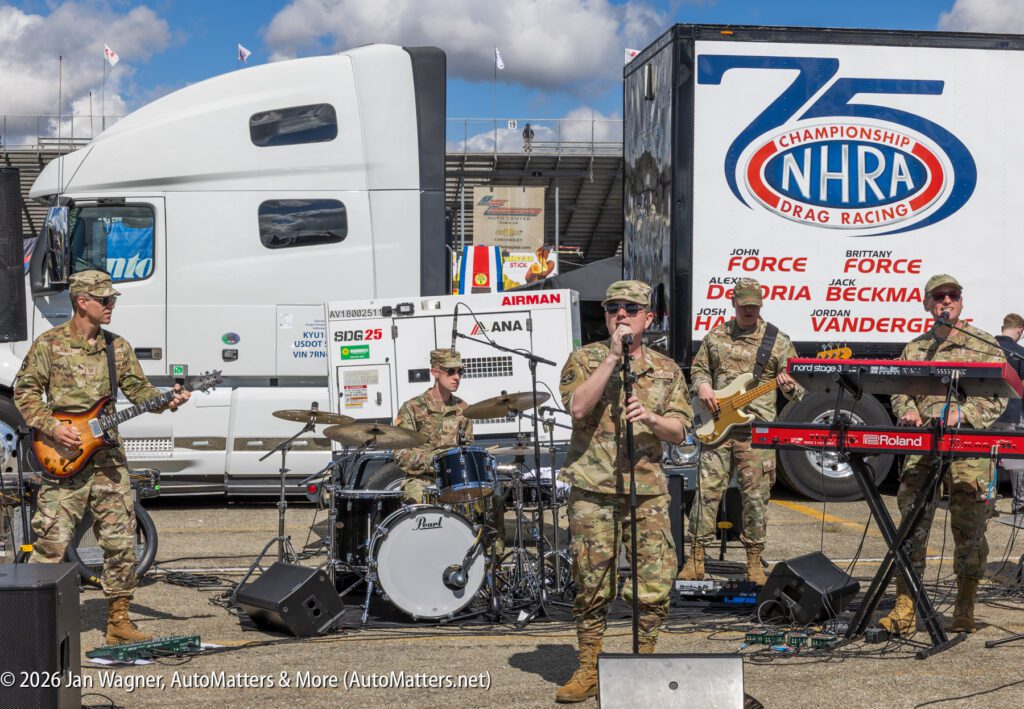 A military band performs outdoors in front of trucks and an NHRA trailer, with members playing guitar, drums, saxophone, keyboard, and singing.