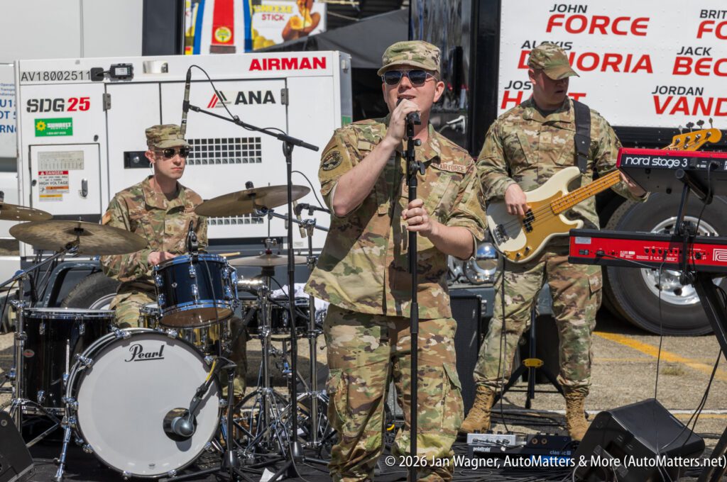 A band of four U.S. military personnel in camouflage uniforms performs outdoors with drums, keyboard, guitar, and a vocalist using a microphone.