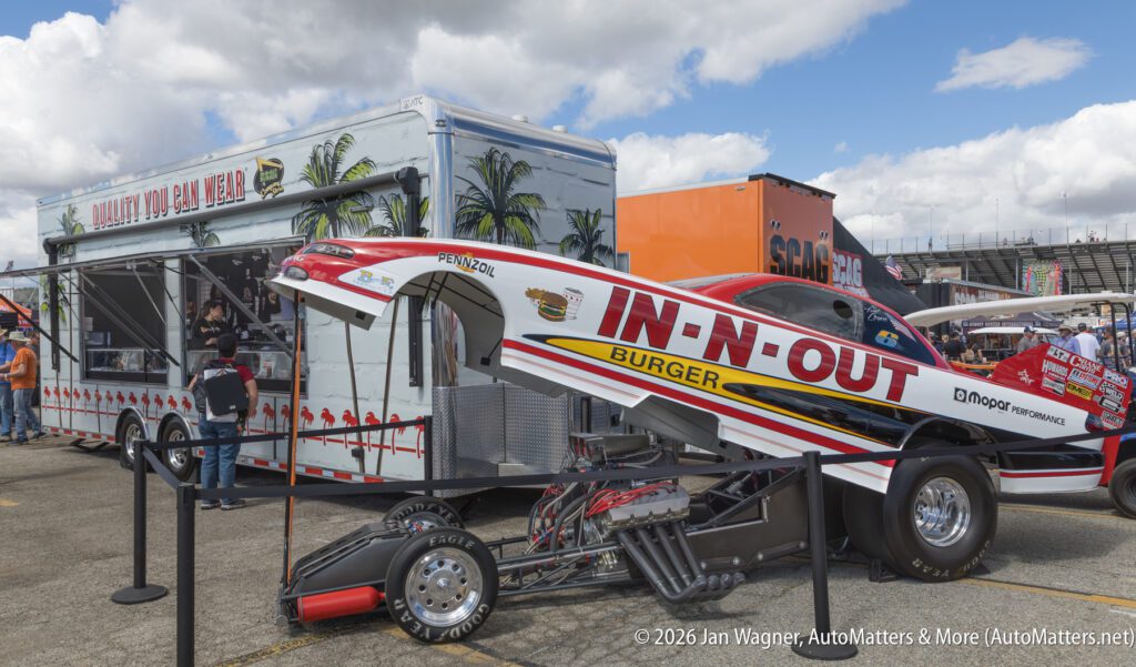 An In-N-Out Burger-themed drag racing car with its body lifted is displayed next to a branded trailer at an outdoor event.