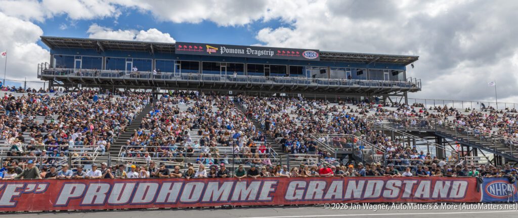 A large crowd sits in the Prudhomme Grandstand at a racing event, with a press box labeled “Pomona Dragstrip” above them under a partly cloudy sky.