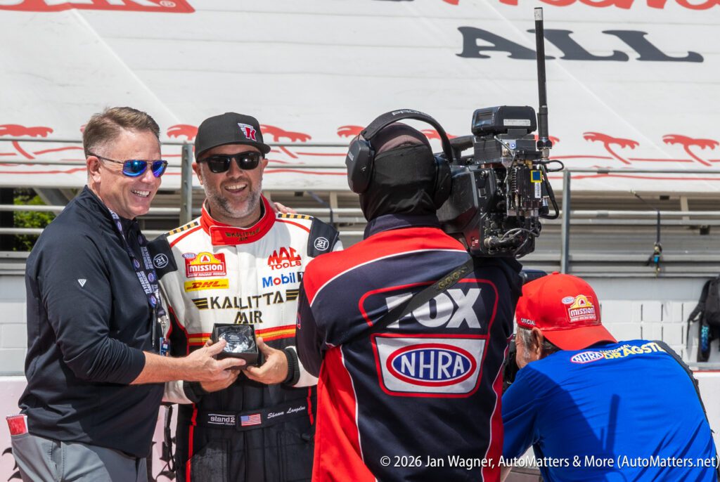 A race car driver poses with an award next to a man, while a TV crew films the scene at a racing event.