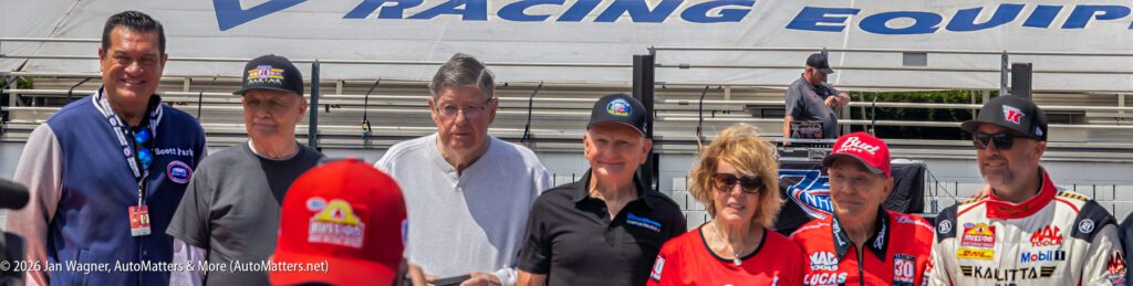 A group of eight people, including racing team members and officials, stand outdoors in front of a racing equipment sign.