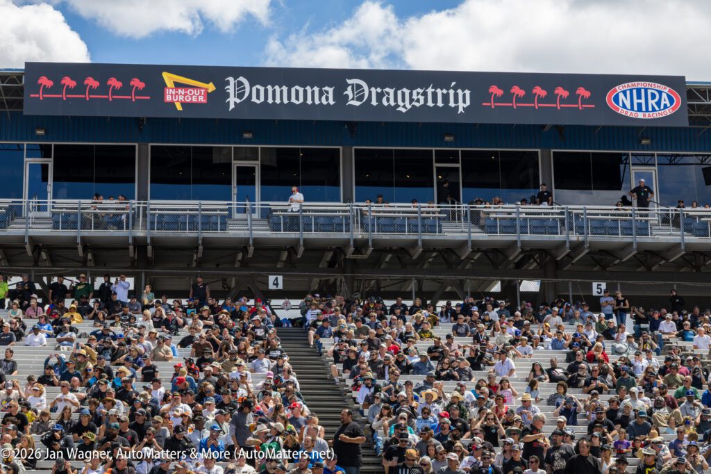 A large crowd sits in the stands beneath the "Pomona Dragstrip" sign at an outdoor racing event on a partly cloudy day.
