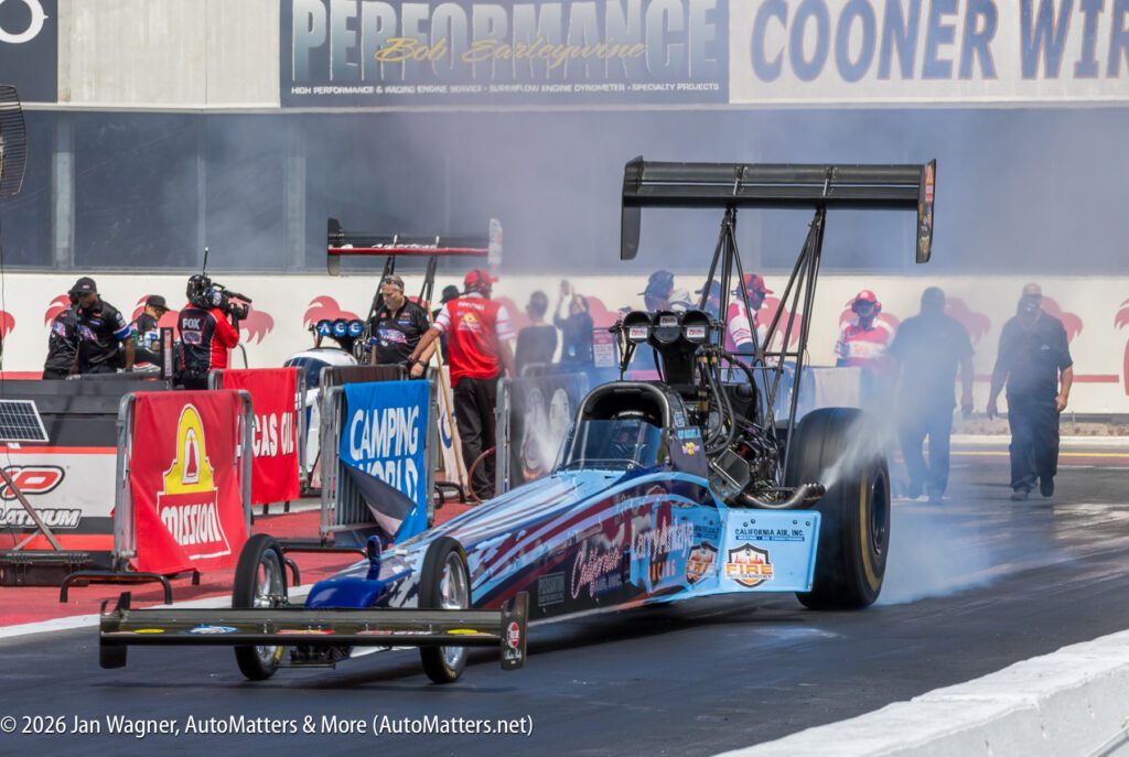 A drag racing car does a burnout at a racetrack, with crew members and officials standing nearby, preparing for a race.