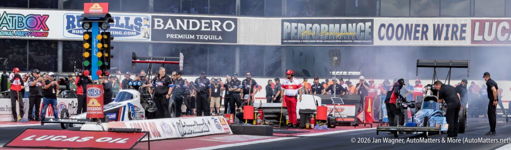 Two drag racing cars prepare to launch at the starting line, surrounded by crew members and spectators at a racetrack.