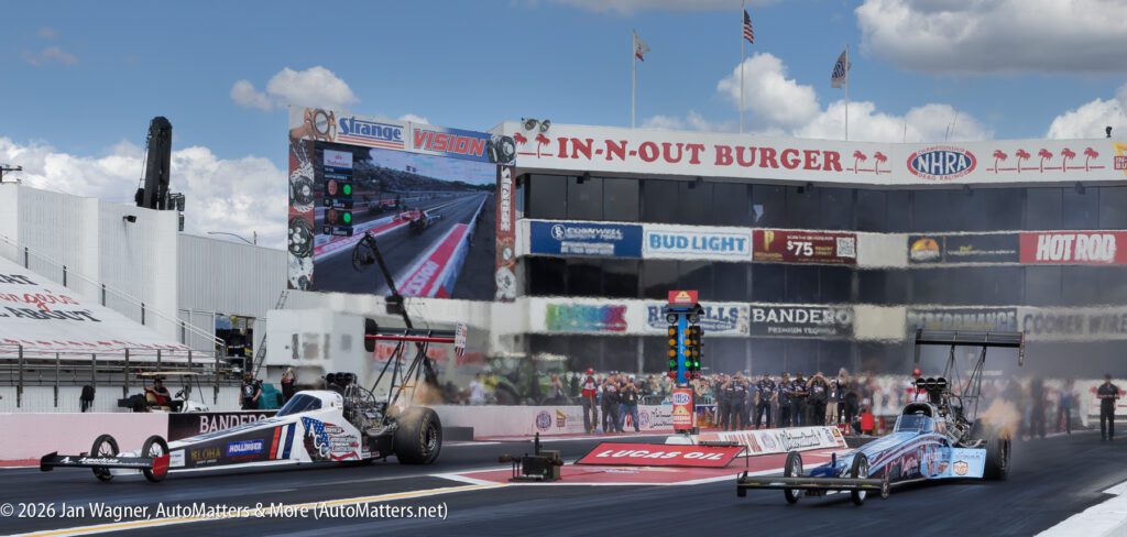 Two drag racing cars launch from the starting line at an NHRA event, with a scoreboard, sponsor signs, and spectators visible in the background.