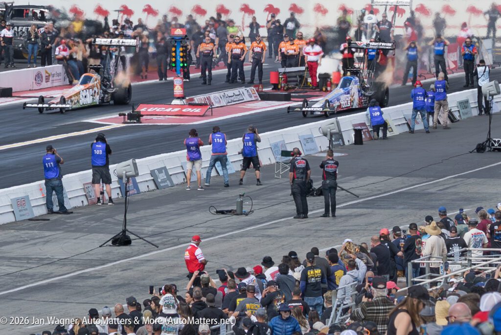 Two drag racing cars line up at the starting line of a racetrack as crew members and photographers stand nearby, with spectators watching from the stands.