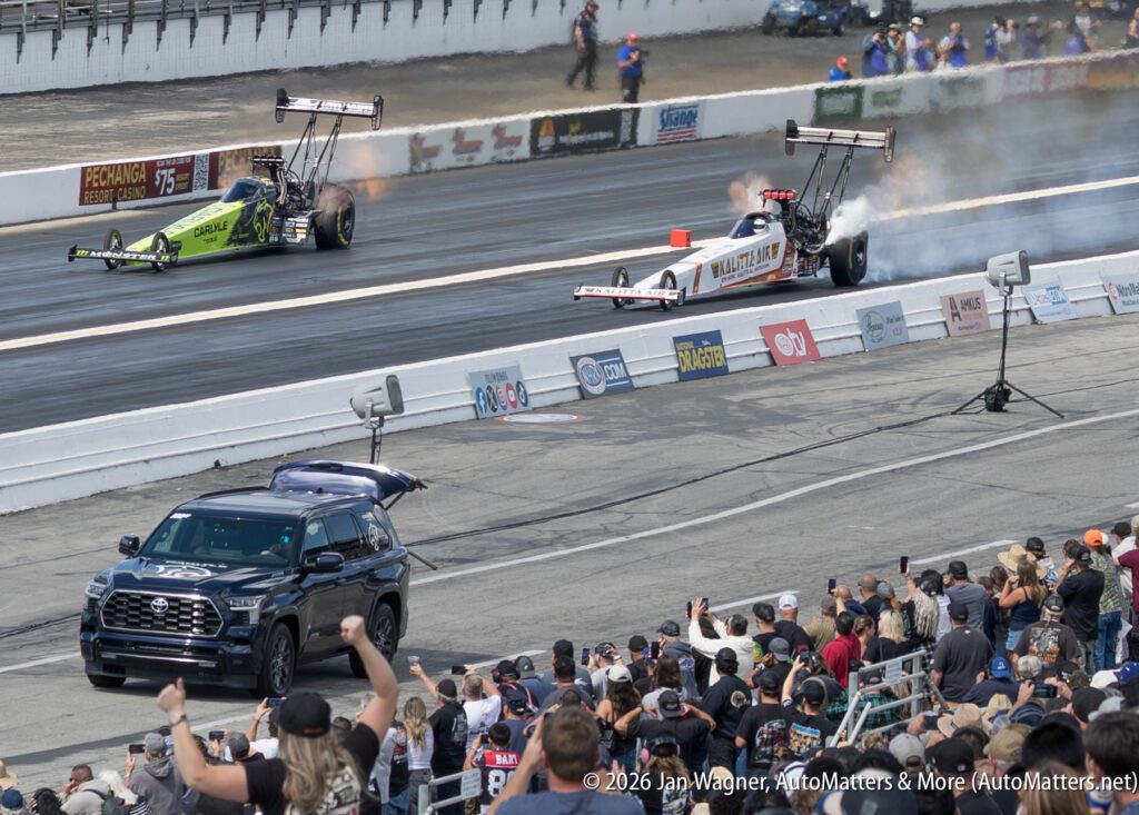 Two drag race cars speed down a track while smoke rises from their tires; a black SUV drives alongside; spectators watch and cheer in the foreground.