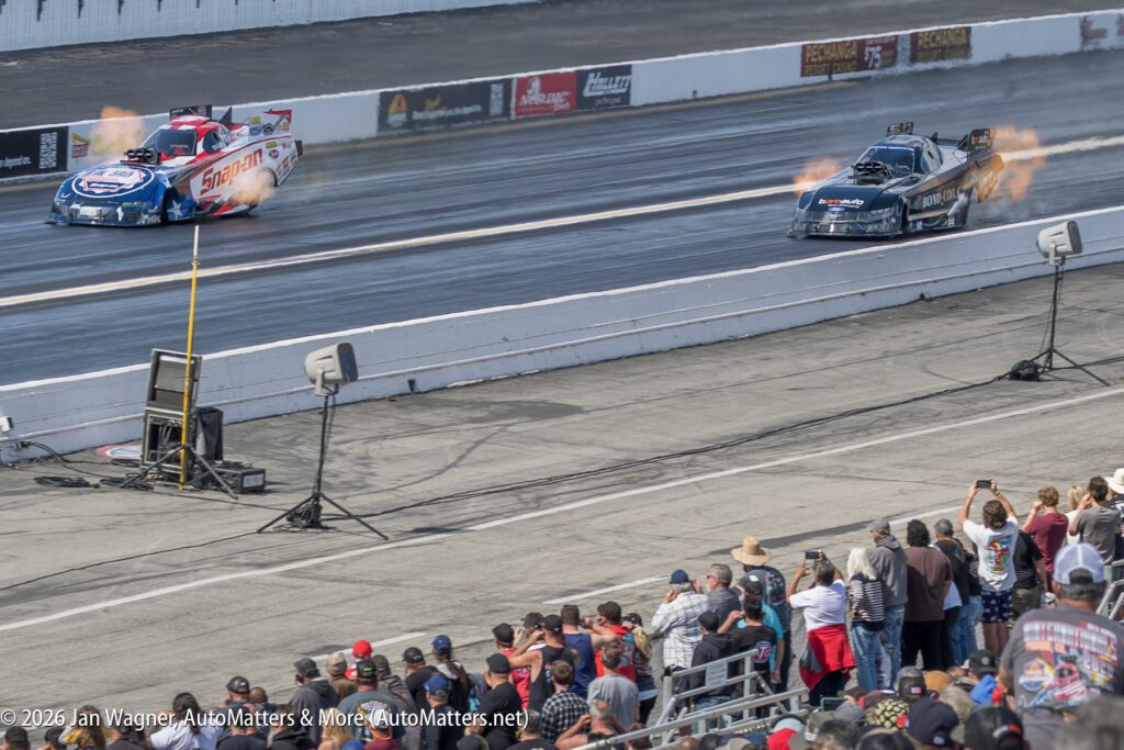 Two drag racing cars speed down a track, flames visible from their exhausts, as spectators watch from the stands.